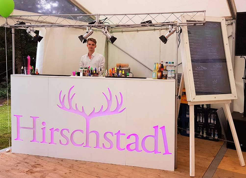 weiße mobile Bar mit Barkeeper. Die Front der Bar ist mit einem pinken Hirschstadlogo versehen.