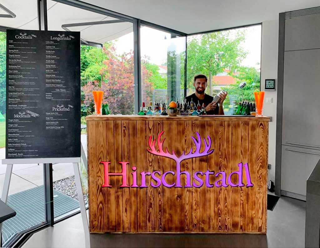 mobile Cocktailbar von der Firma Hirschstadl in geflammter Holzoptik. Auf der mobilen Bar stehen Flaschen und Dekoration. Neben der mobilen Cocktailbar steht eine große Getränkekarte. Hinter der mobilen Cocktailbar steht der Barkeper Chris mit Shaker in der Hand